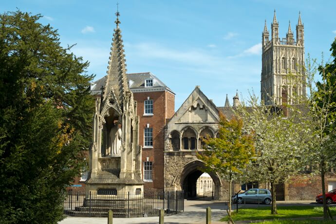 A historic square in Gloucester with a medieval cross monument and the towers of Gloucester Cathedral in the background.