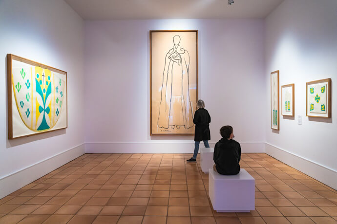 A bright white-walled gallery room with terracotta floors displaying Henri Matisse artworks, including abstract botanical designs and a tall minimalist line drawing, with visitors admiring the art.