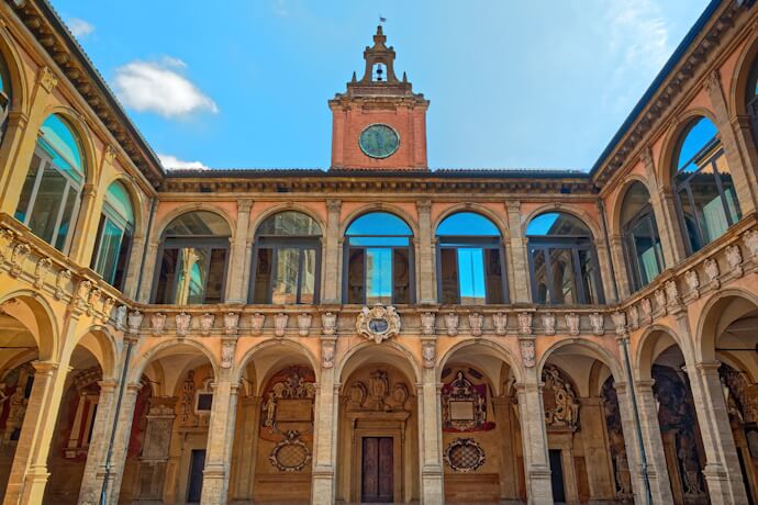 The inner courtyard of the Archiginnasio of Bologna, Emilia-Romagna, Italy, showcasing elegant arched walkways, a central doorway with sculpted decorations, and a clock tower rising above, all in warm terracotta tones under a clear blue sky.