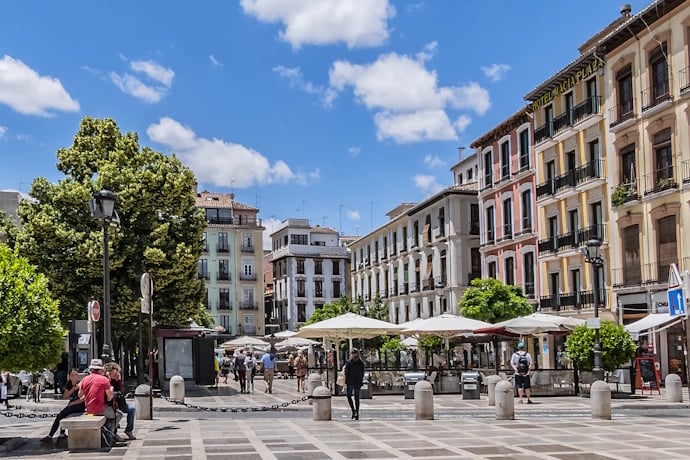 Charming open square in Granada, Spain, called Plaza Nueva, surrounded by traditional Andalusian buildings and cafes with a checkered pavement and trees offering shaded seating.