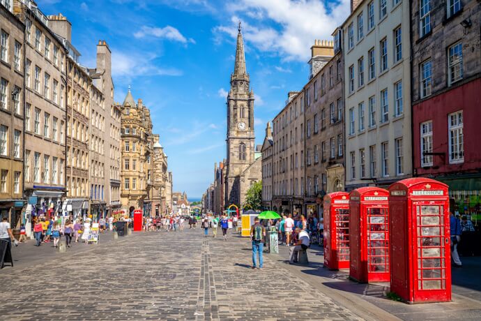 A lively street scene on Edinburgh’s Royal Mile with historic stone buildings, red phone booths, and crowds exploring under a blue sky.