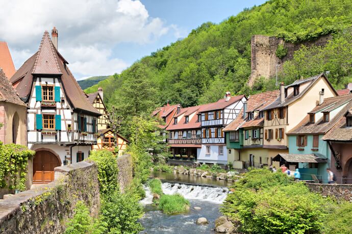 Half-timbered houses lining the river in Kaysersberg, Alsace, France, with medieval architecture and green hills in the background