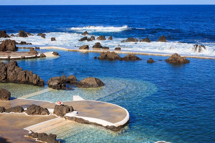 Natural volcanic lava pools filled with clear seawater in Porto Moniz, Madeira, with rugged black rocks and ocean waves crashing beyond.
