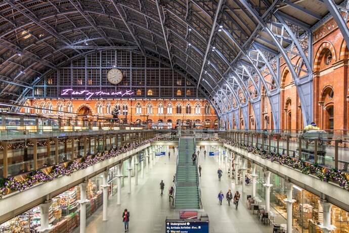 Interior view of St Pancras International Station in London, featuring the arched glass roof, a large clock, festive decorations, and people walking through the main concourse.