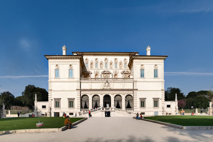The magnificent Villa Borghese with its ornate façade featuring multiple windows, intricate sculptural details and a central section with a grand arched entranceway and a double staircase. In front of the villa, a wide gravel path leads to the stairs, flanked by neatly manicured green lawns.