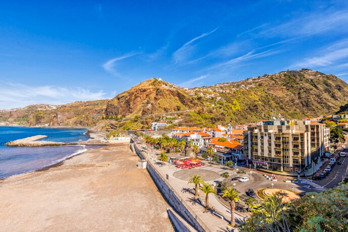 Seaside promenade in Ribeira Brava, Madeira, with palm trees, beachside cafes, and scenic coastal hills under a sunny blue sky.