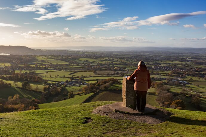 Person standing at a viewpoint overlooking the scenic countryside of the Cotswolds in England, with rolling green fields stretching into the distance.