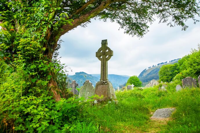 An ancient stone Celtic cross in a serene graveyard surrounded by green hills and trees.