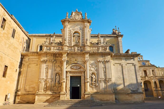 The ornate, light-colored façade of the Basilica di Santa Croce in Lecce, Italy, stands tall against a clear blue sky. The architecture is rich with intricate carvings, sculptures, and decorative elements, showcasing the Baroque style.