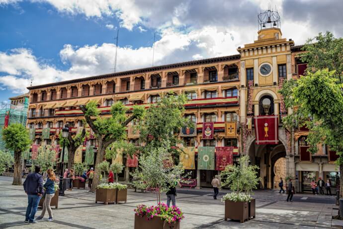 Colorful and festive Plaza de Zocodover in Toledo, Spain, adorned with medieval banners, trees in planters, and visitors walking along the historic arcaded buildings.