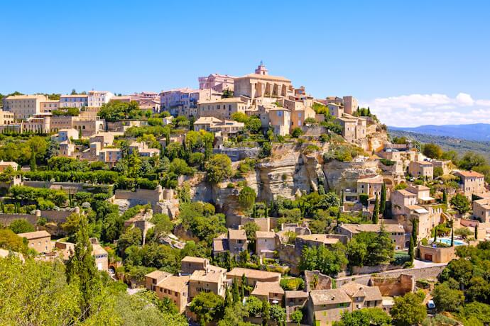 Charming hilltop village of Gordes in Provence, France, with its stone houses nestled into the cliffs, surrounded by lush greenery under a bright blue summer sky.
