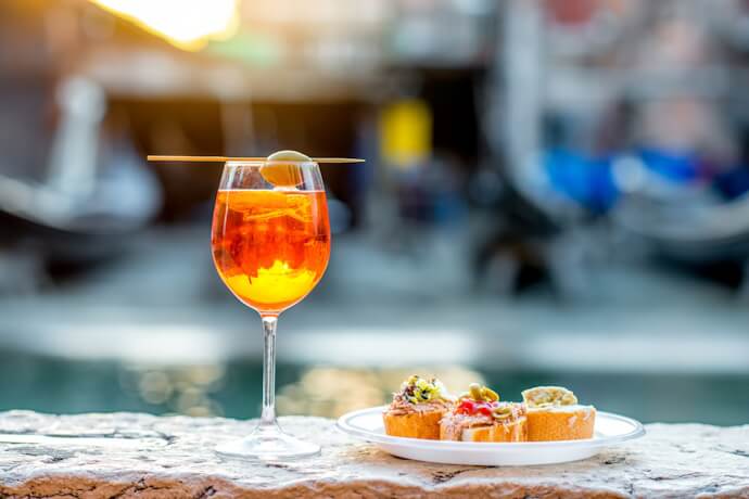 Close-up of a vibrant orange Aperol Spritz with an olive garnish and a plate of Venetian cicchetti on a stone ledge, with gondolas softly blurred in the background.
