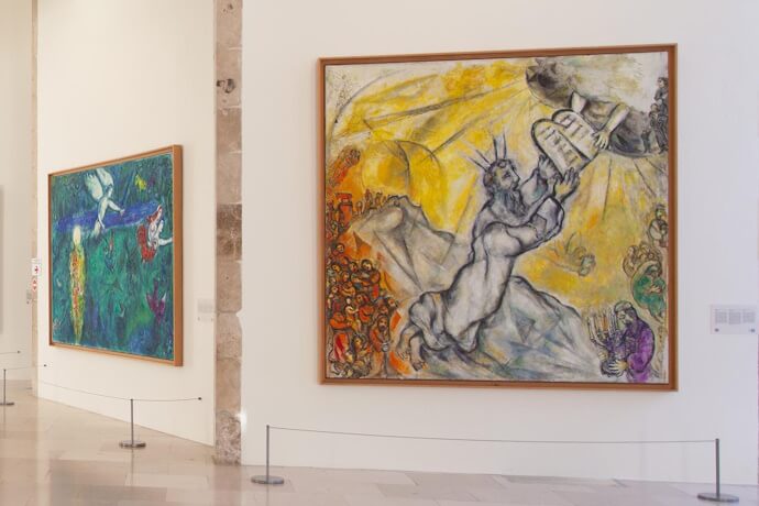 A spacious gallery room displaying large Marc Chagall paintings filled with vivid colors and biblical themes, with protective barriers in front of the artworks.