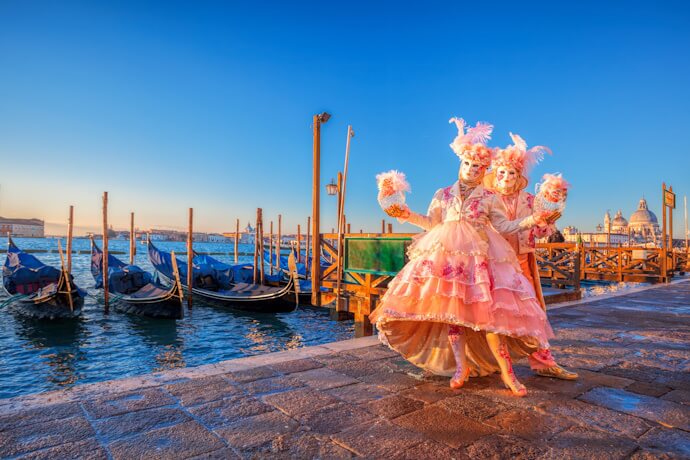 Two elaborately costumed and masked figures in pink dresses pose at sunrise during Venice Carnival, with gondolas moored along the lagoon and clear blue skies.