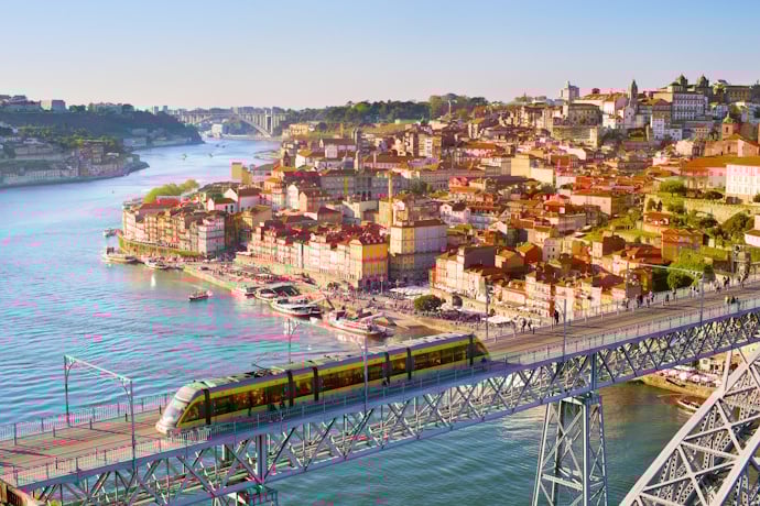 A scenic view of Porto, Portugal, with a yellow metro train crossing the iconic Dom Luís I Bridge above the Douro River and the historic Ribeira district in the background.
