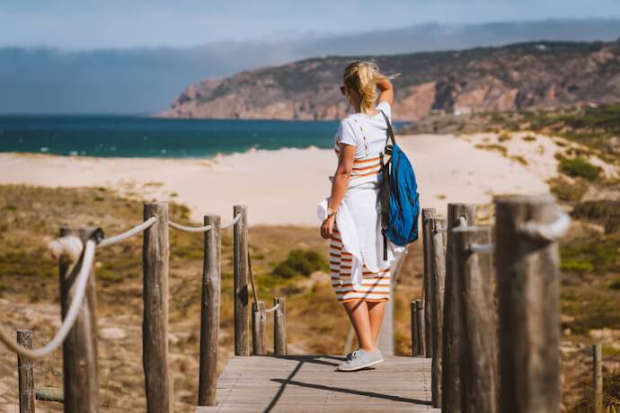Woman walking along a wooden boardwalk toward Guincho Beach in Portugal with cliffs and turquoise sea in the background.