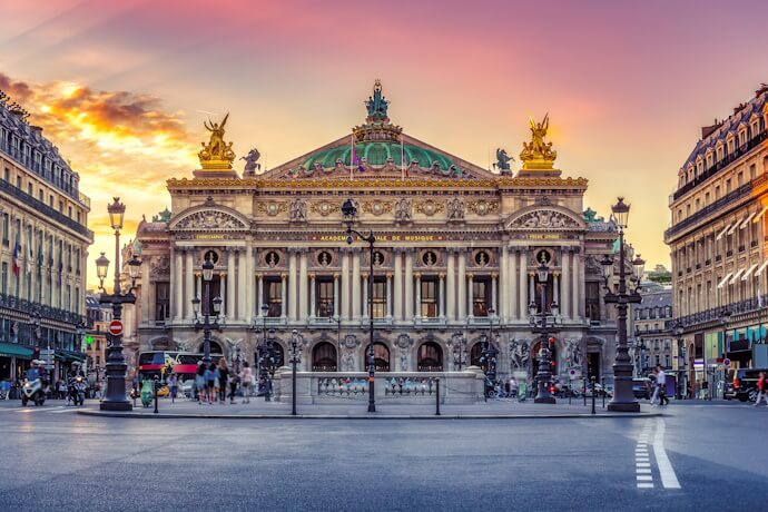 The opulent façade of the Palais Garnier in Paris illuminated at sunset, with golden statues, grand columns, and people gathered at the entrance.