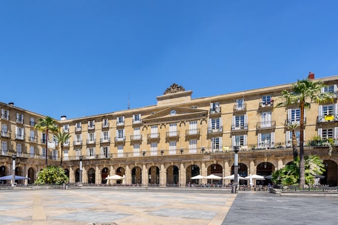 Spacious arcaded square in Bilbao, Spain, known as Plaza Nueva, with neoclassical architecture, palm trees, and outdoor seating areas around the central empty courtyard.