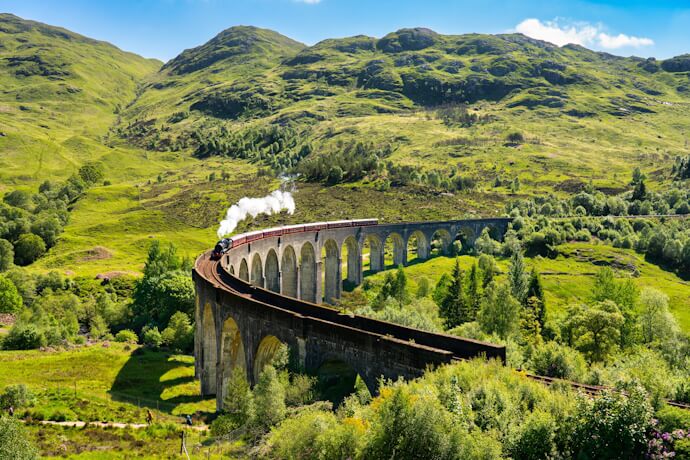 Steam train crossing the Glenfinnan Viaduct amid the green Highlands landscape in Scotland.