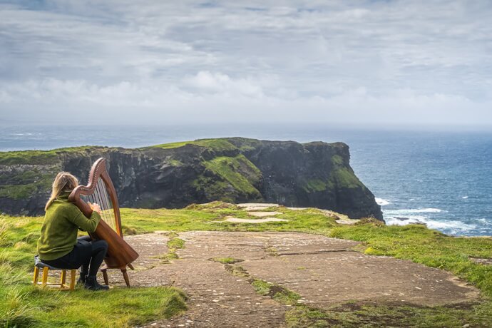 A musician playing a harp atop sea cliffs, with waves crashing below.