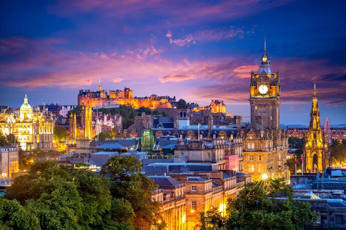 The city of Edinburgh illuminated at dusk, featuring the Balmoral Clock Tower and Edinburgh Castle glowing under a colorful sky.