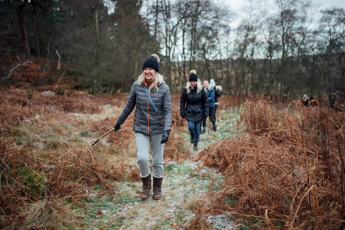 Group of people dressed in winter clothing hiking through a frosty woodland trail, with dry ferns and bare trees surrounding the path.