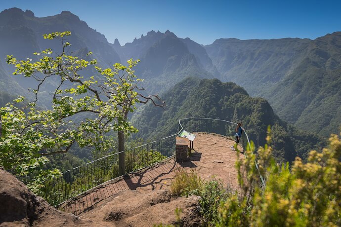 Scenic viewpoint along the Levada do Balcões trail in Ribeiro Frio, Madeira, with a person standing on a rocky terrace overlooking dramatic green mountain peaks under a clear blue sky.