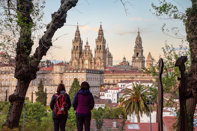 Pilgrims viewing the towering spires of Santiago de Compostela Cathedral at sunset, framed by trees and city rooftops.