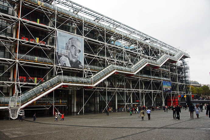 Exterior of the Centre Pompidou in Paris, showcasing its high-tech architecture with exposed pipes and escalators, and visitors in the open plaza.