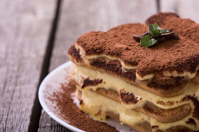 A close-up of creamy tiramisu dusted with cocoa powder, garnished with mint and chocolate on a rustic wooden table.