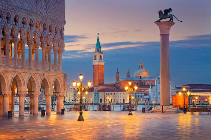 Early morning view of the Piazzetta San Marco in Venice with warm streetlights glowing, Doge’s Palace arches on the left, the Lion of Saint Mark column, and the Church of San Giorgio Maggiore across the lagoon under a soft dawn sky.