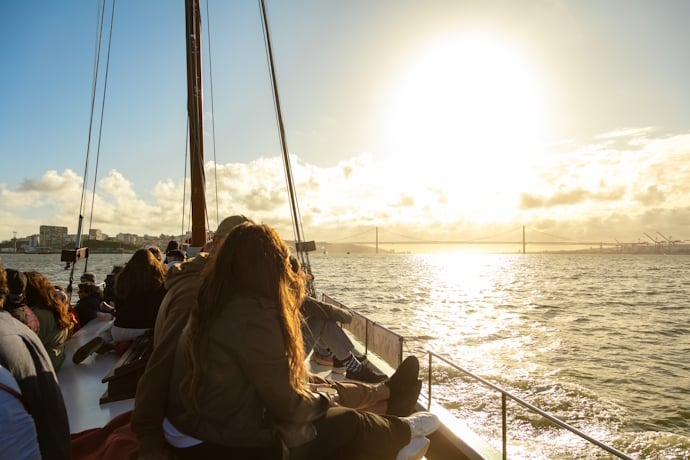 Passengers relaxing on a sailboat at sunset with the 25 de Abril Bridge silhouetted on the horizon.