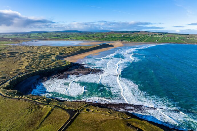 Aerial view of rolling waves hitting a sandy beach with patchwork farmland beyond.