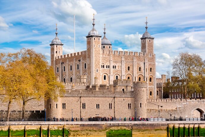 The historic Tower of London fortress with its stone walls, turrets, and moat, surrounded by visitors on a sunny day.