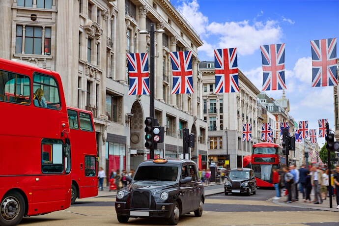 Busy London street scene with red double-decker buses, a black taxi, and Union Jack flags hanging above the road, set against historic buildings and a bright blue sky.
