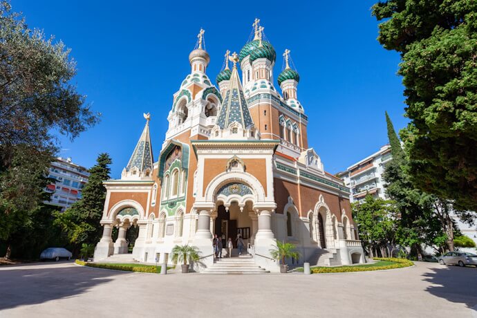 The striking Russian Orthodox Cathedral in Nice featuring ornate brickwork, green onion domes with gilded details, and arched entrances, surrounded by trees and visitors.