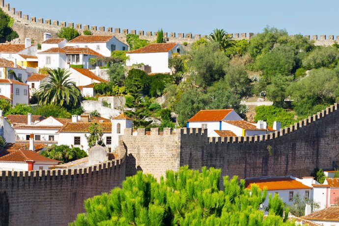 A view of the medieval walled town of Óbidos, Portugal, with whitewashed houses, terracotta roofs, and olive trees framed by the town’s crenelated stone fortifications.