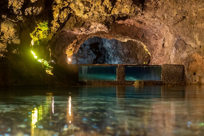 Illuminated volcanic cave interior in São Vicente, Madeira, featuring clear water pools and textured lava rock walls with ambient lighting.