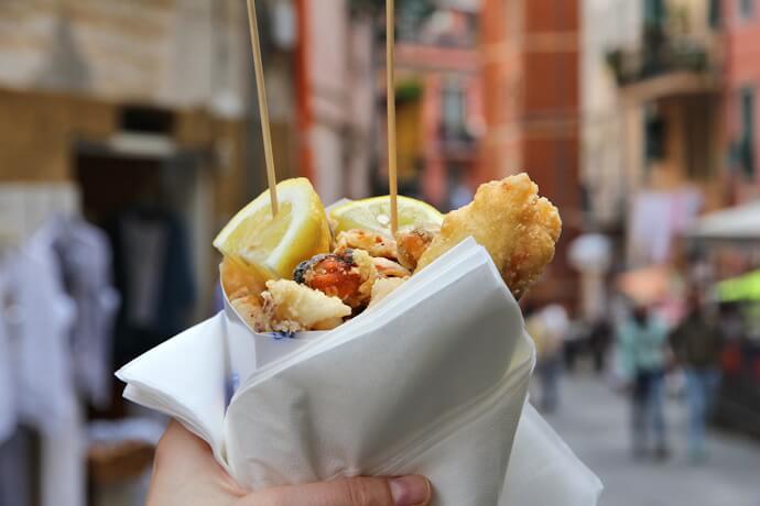 Hand holding a paper cone filled with fried seafood and lemon slices on a street in Italy.