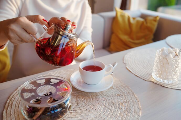 Close-up of a person pouring tea from a glass teapot into a white teacup at a cozy table setting with placemats, cushions, and a jar of sugar cubes.