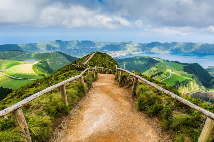 A winding trail leads along a green volcanic ridge in the Azores, opening onto sweeping views of crater lakes and rolling hills.