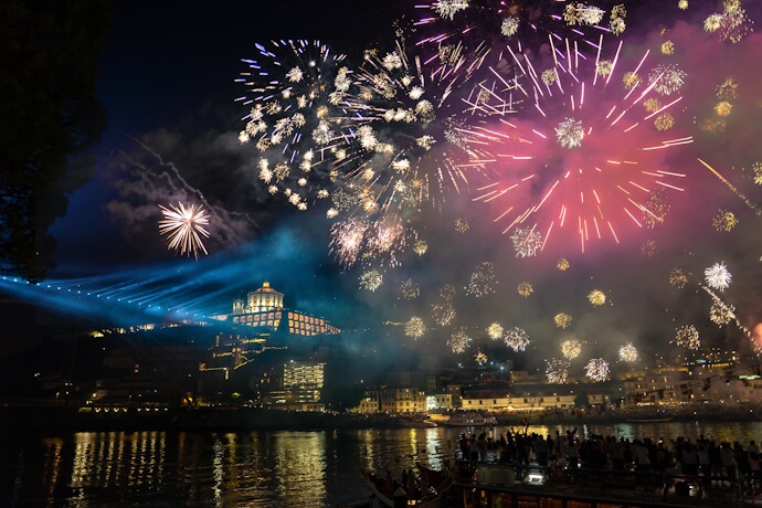 Night falls over Porto, as fireworks burst above the Douro River, illuminating the city skyline and reflecting off the water during a festive celebration.
