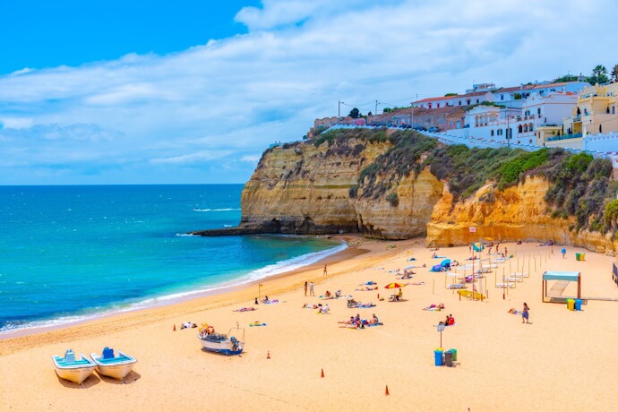 Sunlight reflects off golden sand and turquoise waters in Carvoeiro, Algarve, with dramatic cliffs overlooking a relaxed beach scene.