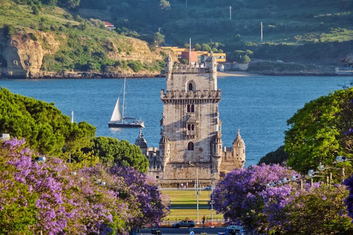 Belém Tower framed by jacaranda blooms along the Tagus River, Lisbon.