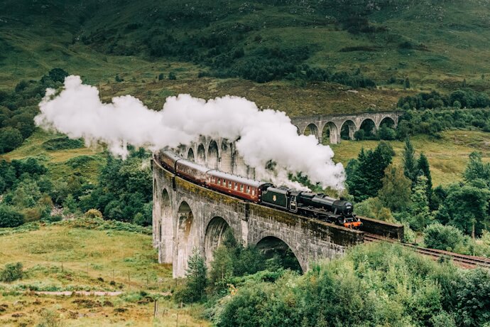 Steam train crossing the Glenfinnan Viaduct, surrounded by the dramatic green landscape of the Scottish Highlands.