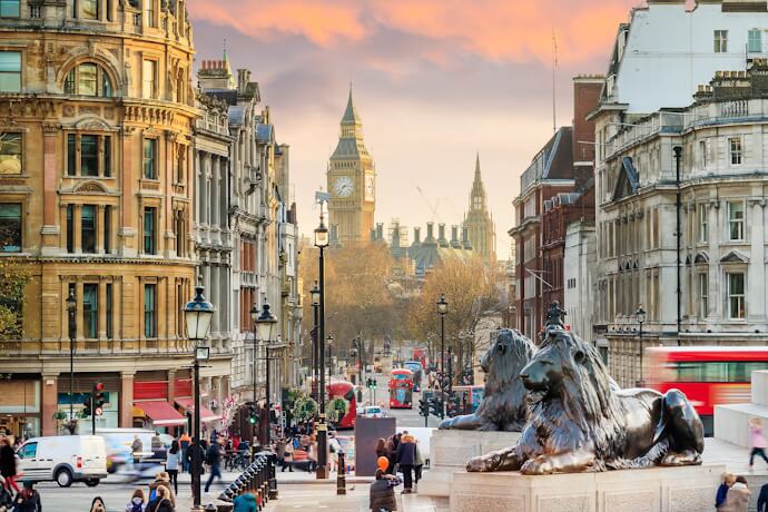 View of Trafalgar Square in London, with the iconic lion statues in the foreground and Big Ben visible in the background among historic buildings.