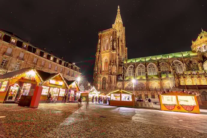 Night view of Strasbourg Cathedral towering over festive Christmas market chalets glowing below.