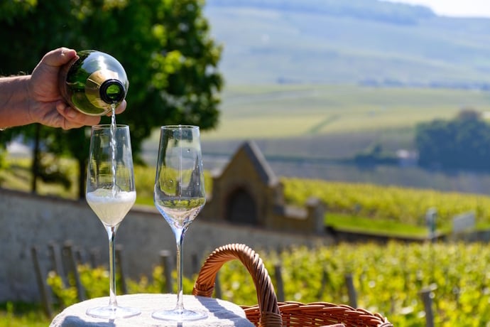 A bottle of Champagne is poured into flutes with sunlit vineyards and rolling hills in the background.