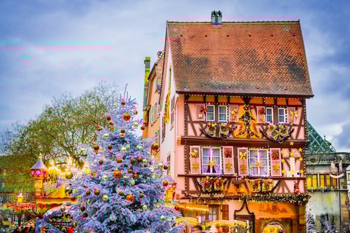 A half-timbered house dressed in holiday garlands stands beside a snow-kissed Christmas tree at the market, in Colmar