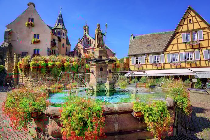 The village square in Eguisheim blooms with flowers around a central fountain, surrounded by storybook Alsatian houses.